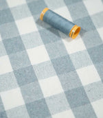 Angled view of Aldsworth Check Haze checked fabric with a coordinating blue thread spool resting on the textured blue and white woven surface.