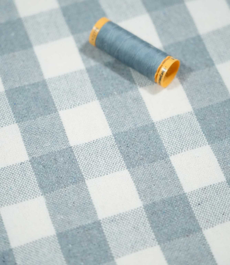 Angled view of Aldsworth Check Haze checked fabric with a coordinating blue thread spool resting on the textured blue and white woven surface.