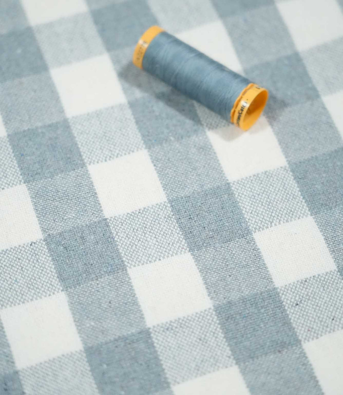 Angled view of Aldsworth Check Haze checked fabric with a coordinating blue thread spool resting on the textured blue and white woven surface.