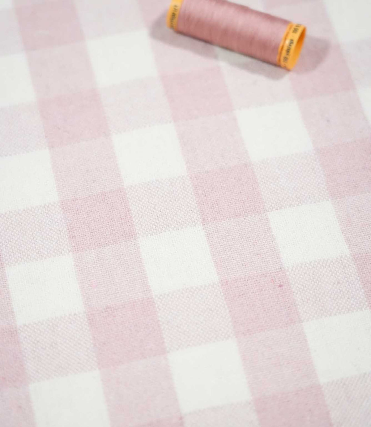 Angled view of Aldsworth Check Pink fabric with a coordinating pink thread spool resting on the soft pink and white checked woven surface.