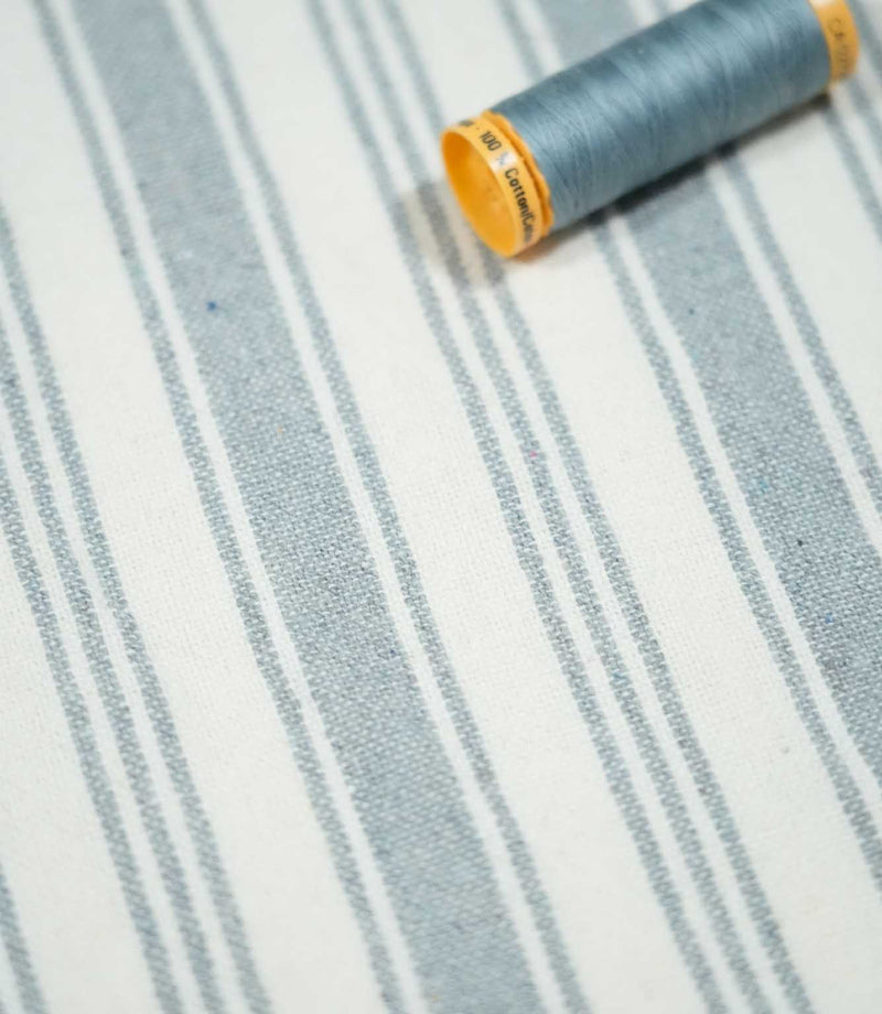 Aldsworth Stripe Haze fabric with a coordinating blue sewing thread spool, displaying its classic striped pattern and woven texture.