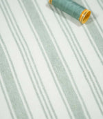 Aldsworth Stripe Mint striped fabric displayed flat with a coordinating green sewing thread spool, showcasing its delicate stripes and woven texture.