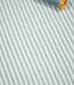 Aldsworth Ticking Haze striped fabric laid flat beside a coordinating blue thread spool, highlighting its narrow stripes and textured weave.