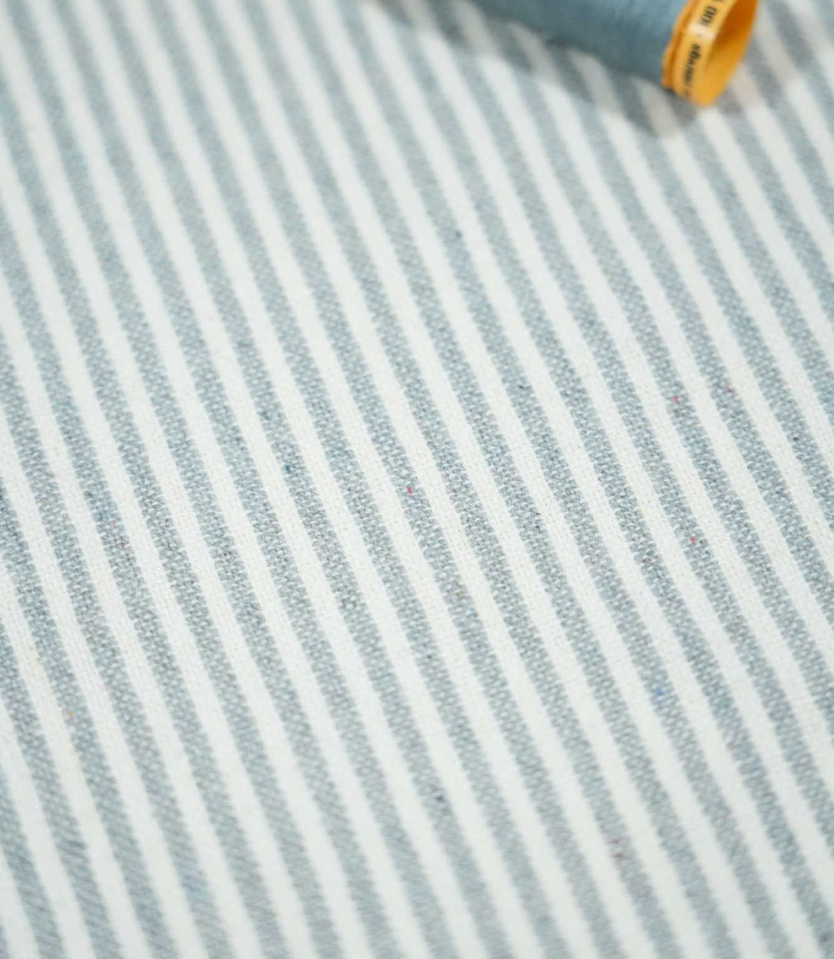 Aldsworth Ticking Haze striped fabric laid flat beside a coordinating blue thread spool, highlighting its narrow stripes and textured weave.