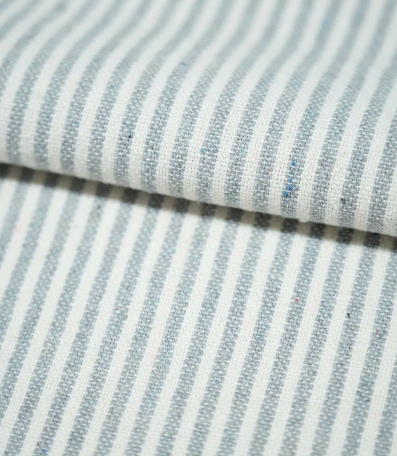 Folded Aldsworth Ticking Haze fabric showing classic blue-grey and white ticking stripes, with visible woven texture and subtle coloured flecks.