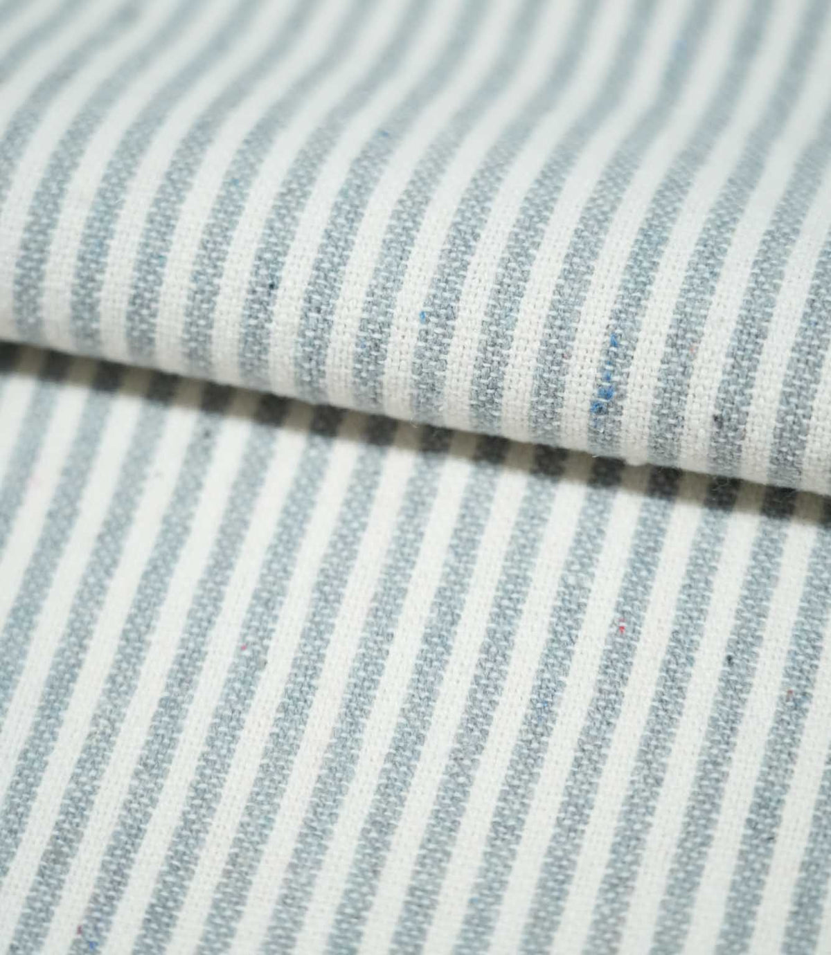 Folded Aldsworth Ticking Haze fabric showing classic blue-grey and white ticking stripes, with visible woven texture and subtle coloured flecks.