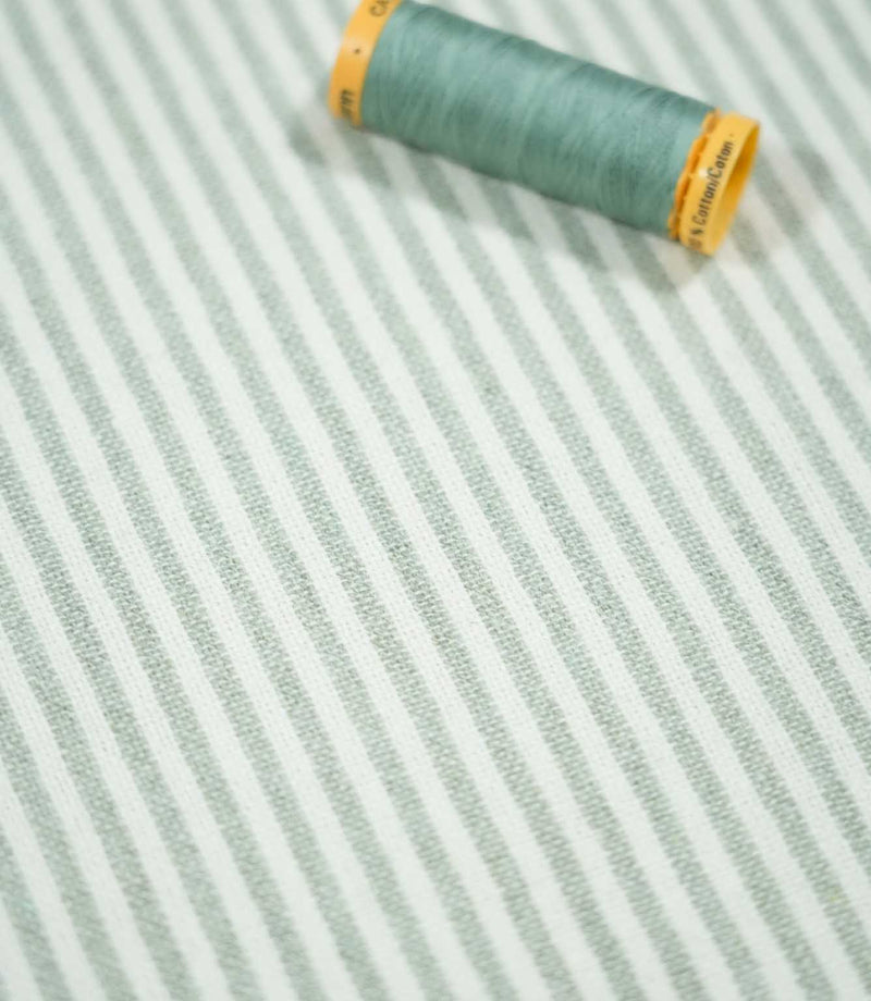 Aldsworth Ticking Mint striped fabric laid flat beside a coordinating green thread spool, highlighting its delicate stripes and textured weave.