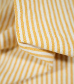Close-up of folded Aldsworth Ticking Ochre fabric highlighting the textured weave, fine ochre stripes and contrasting off-white background.