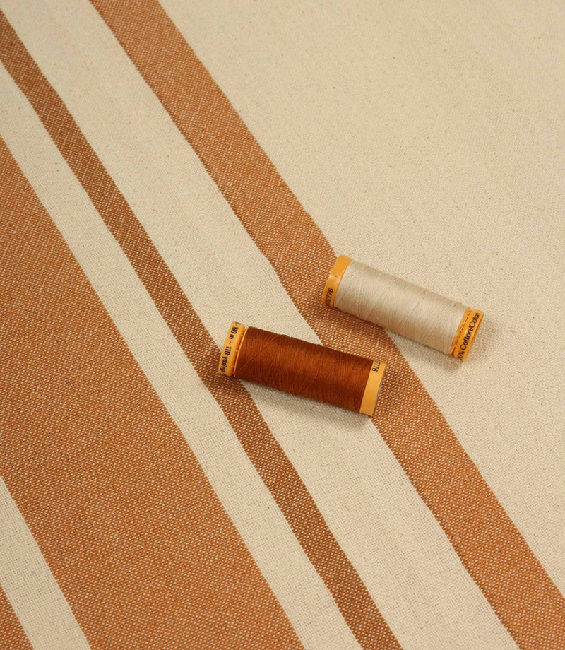 Cream and terracotta striped furnishing fabric displayed with matching thread reels to showcase colour and weave detail.