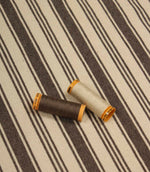 Cream and dark brown striped furnishing fabric displayed with coordinating thread reels to highlight colour matching and weave detail.