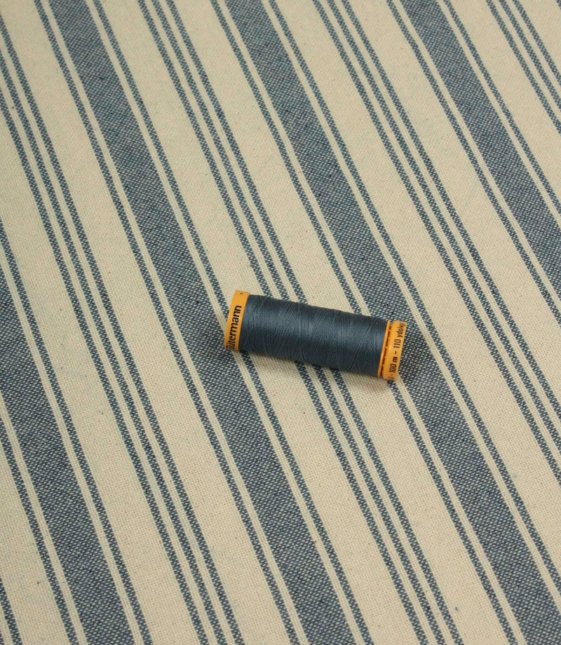 Denim-blue striped fabric laid flat with a coordinating spool of navy thread, highlighting the fabric’s classic ticking stripe design and upholstery weight texture.