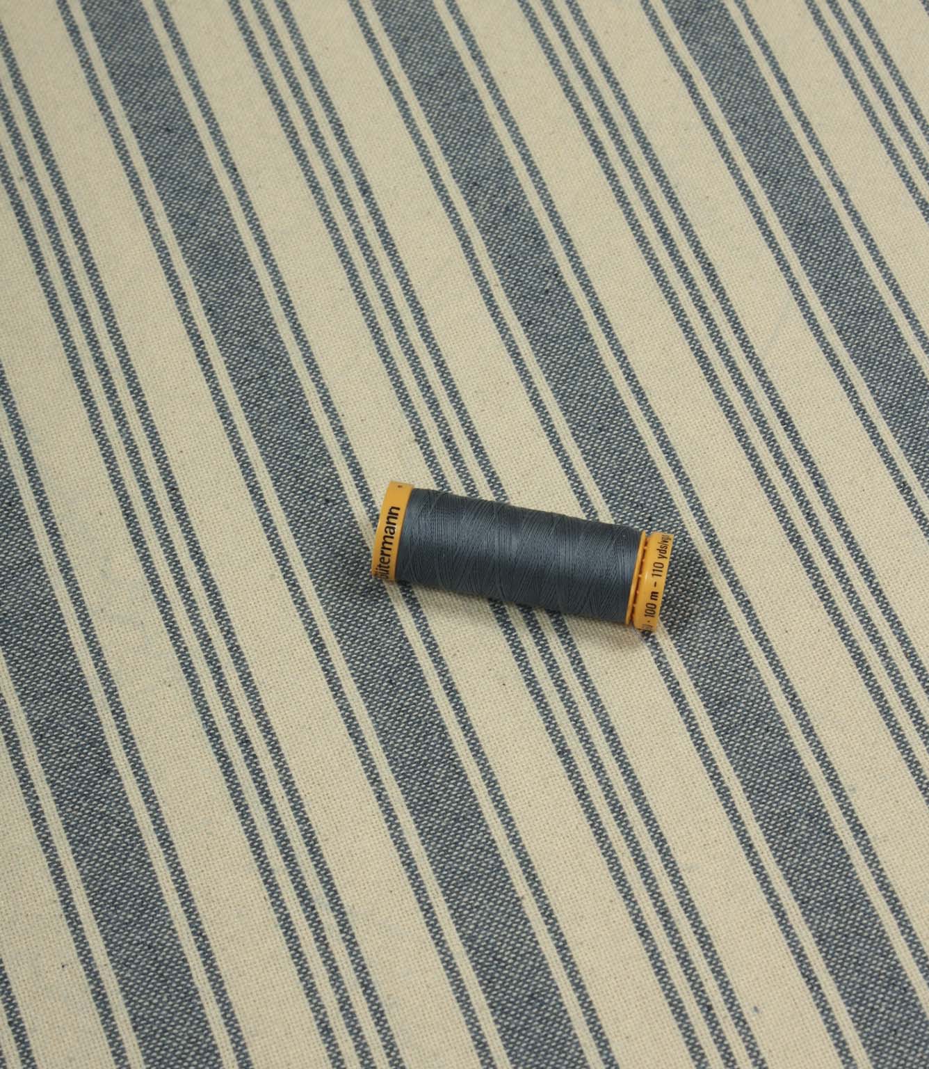 Denim-blue striped fabric laid flat with a coordinating spool of navy thread, highlighting the fabric’s classic ticking stripe design and upholstery weight texture.