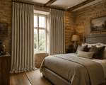 Luxury Cotswold bedroom with blue ticking stripe curtains, exposed stone walls and oak flooring, softly lit by natural daylight. Upholstered bed with layered neutral bedding and plain front cushion creates a warm, timeless countryside hotel aesthetic.