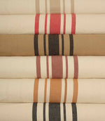 Close-up of rolled striped fabrics highlighting the textured weave and bold stripe patterns in ochre, rose pink, and charcoal against natural cream backgrounds.