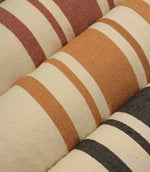Woven striped furnishing fabrics in coral red, camel brown, and neutral beige tones, displayed as rolled fabric bolts with classic wide stripe detailing.
