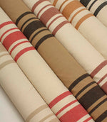 Neutral and earthy striped fabric rolls arranged diagonally, showcasing classic woven stripe designs in beige, coral, camel, ochre, rose, and charcoal colourways.