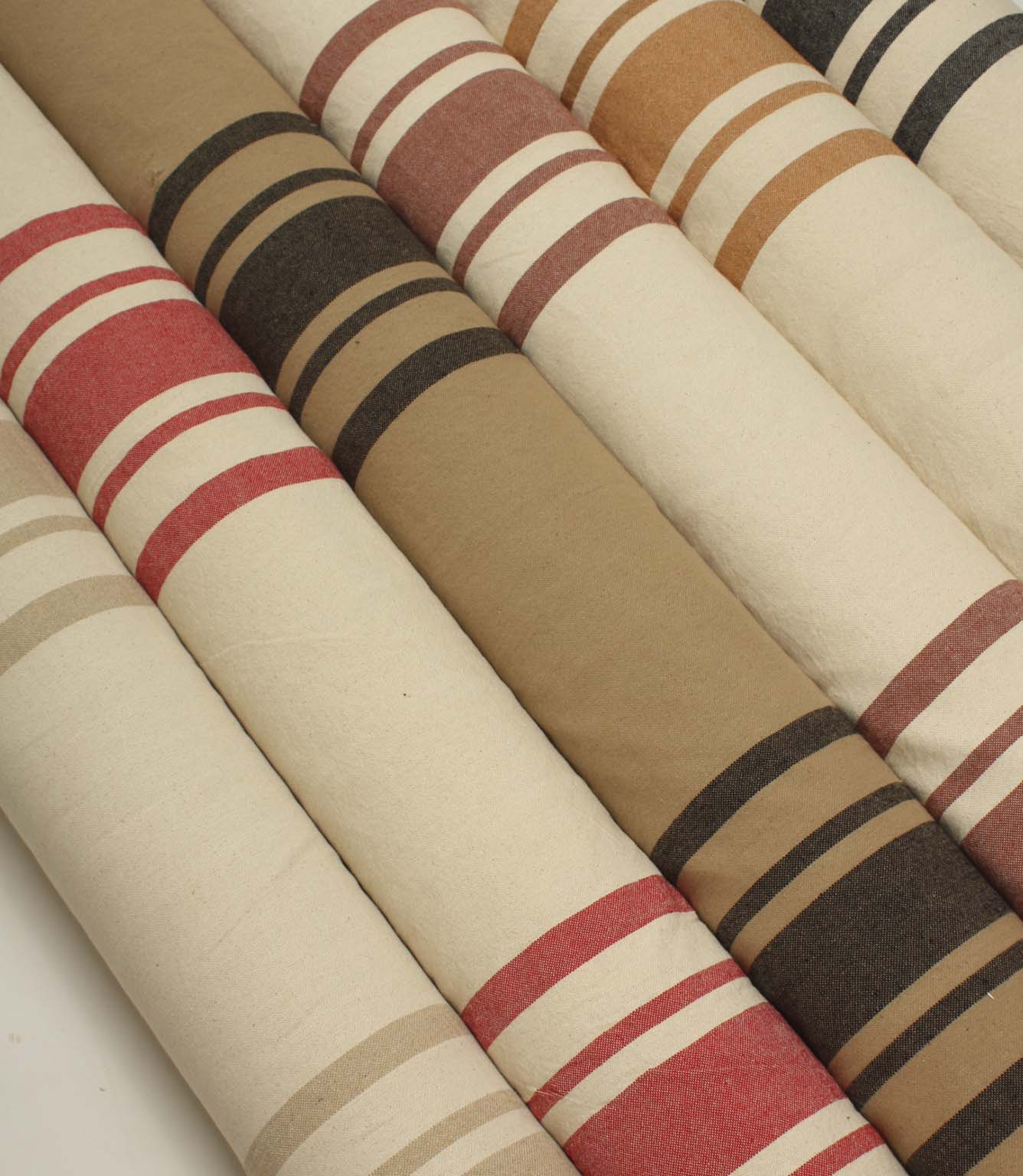 Neutral and earthy striped fabric rolls arranged diagonally, showcasing classic woven stripe designs in beige, coral, camel, ochre, rose, and charcoal colourways.