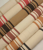 Collection of striped upholstery and curtain fabrics arranged in horizontal rolls, showcasing classic woven stripe designs in soft neutral and earthy tones.