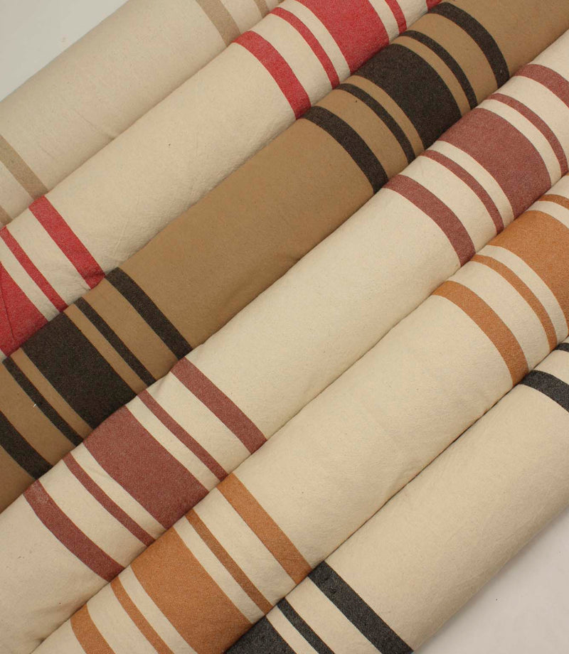 Collection of striped upholstery and curtain fabrics arranged in horizontal rolls, showcasing classic woven stripe designs in soft neutral and earthy tones.