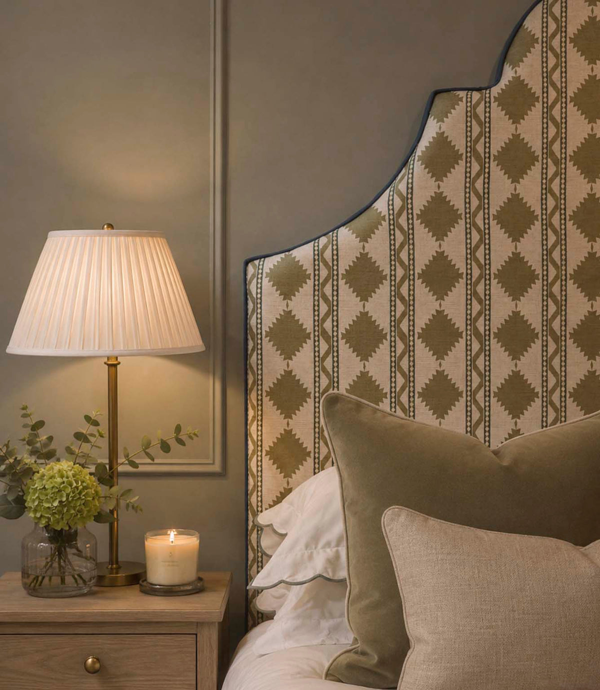 Arlo Geo Olive fabric upholstered headboard featuring repeating olive geometric motifs and striped detailing on a natural background, finished with contrasting blue piping and styled with olive and neutral cushions.