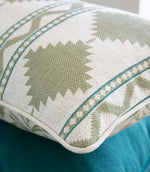 A close-up of a woven throw pillow featuring a sage green and cream geometric pattern with teal accent stripes, resting on a teal surface.