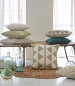 A styled interior scene with multiple decorative throw pillows arranged on a bench and floor, alongside glass bottles and a woven rug in a bright room with curtains.