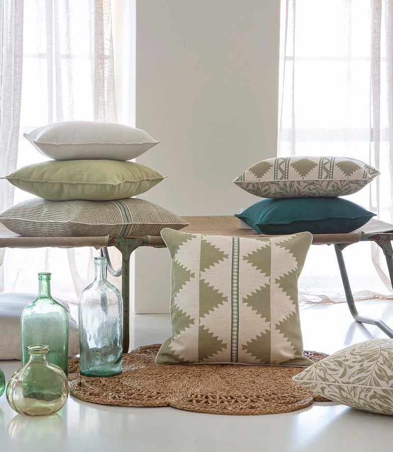 A styled interior scene with multiple decorative throw pillows arranged on a bench and floor, alongside glass bottles and a woven rug in a bright room with curtains.