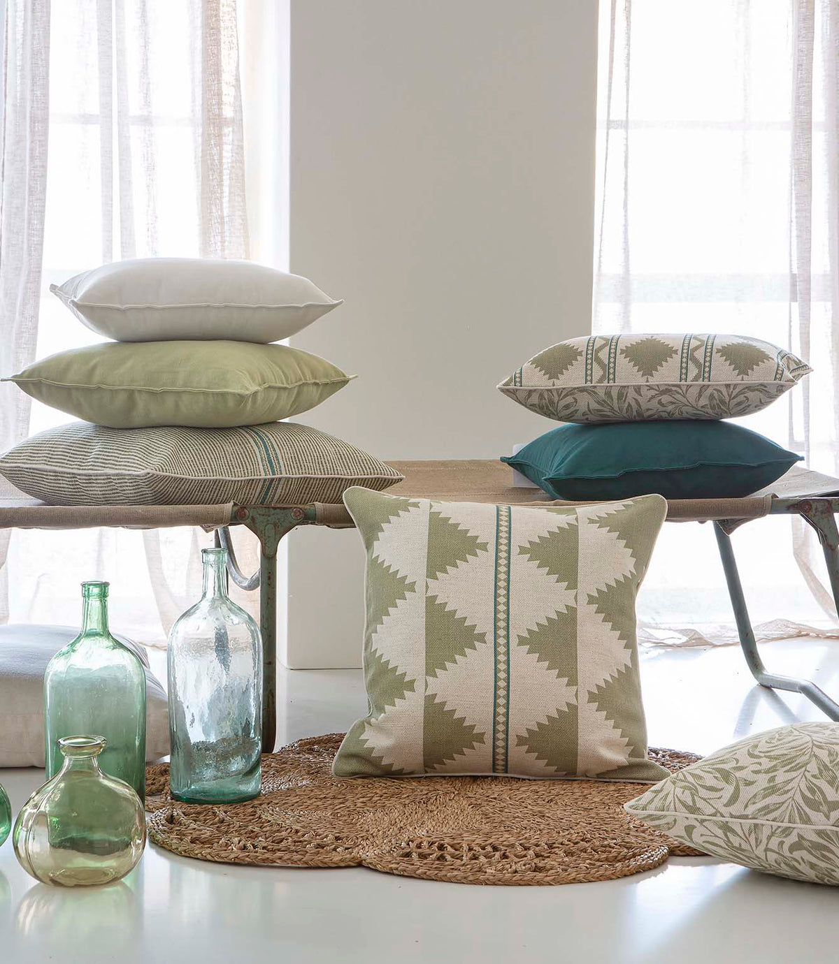 A styled interior scene with multiple decorative throw pillows arranged on a bench and floor, alongside glass bottles and a woven rug in a bright room with curtains.