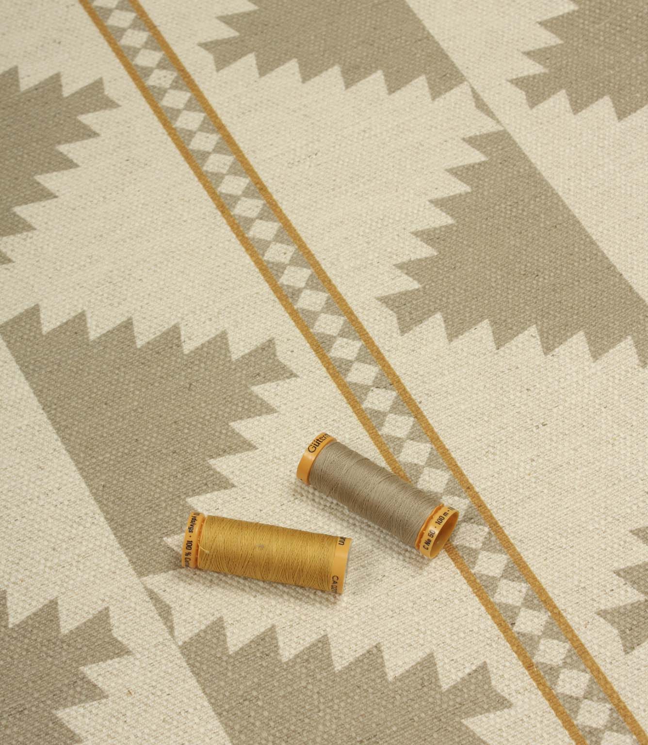 Southwestern-style furnishing fabric displayed with coordinating thread reels, featuring muted taupe diamond motifs on a natural cream woven background.