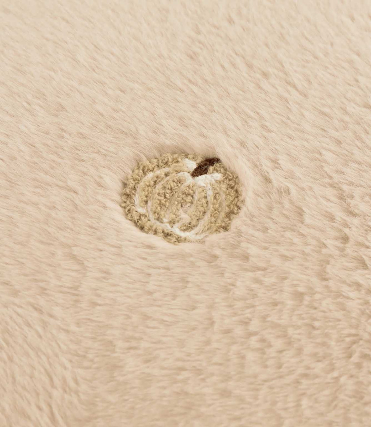Close-up of a beige embroidered pumpkin motif with a brown stem on the soft natural cream faux fur cushion fabric.
