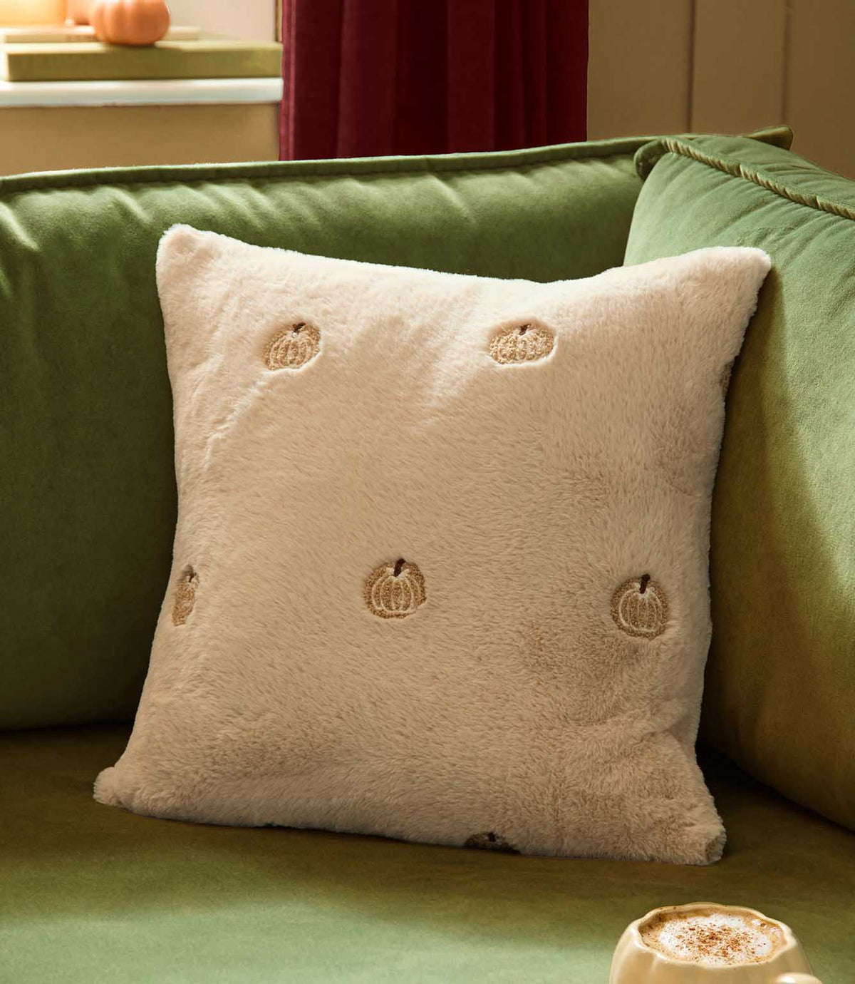 Natural cream Autumn Pumpkins embroidered faux fur cushion styled upright on a green velvet sofa in a cosy autumn living room.