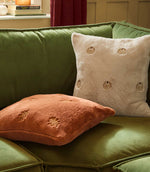Natural cream pumpkin embroidered faux fur cushion styled with a rust orange pumpkin cushion on a green velvet sofa in an autumnal interior.