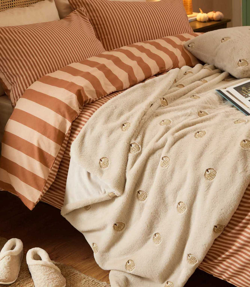 Autumn bedroom styled with the natural cream embroidered pumpkin faux fur throw layered over rust and cream striped bedding, with a matching cushion and cosy slippers.