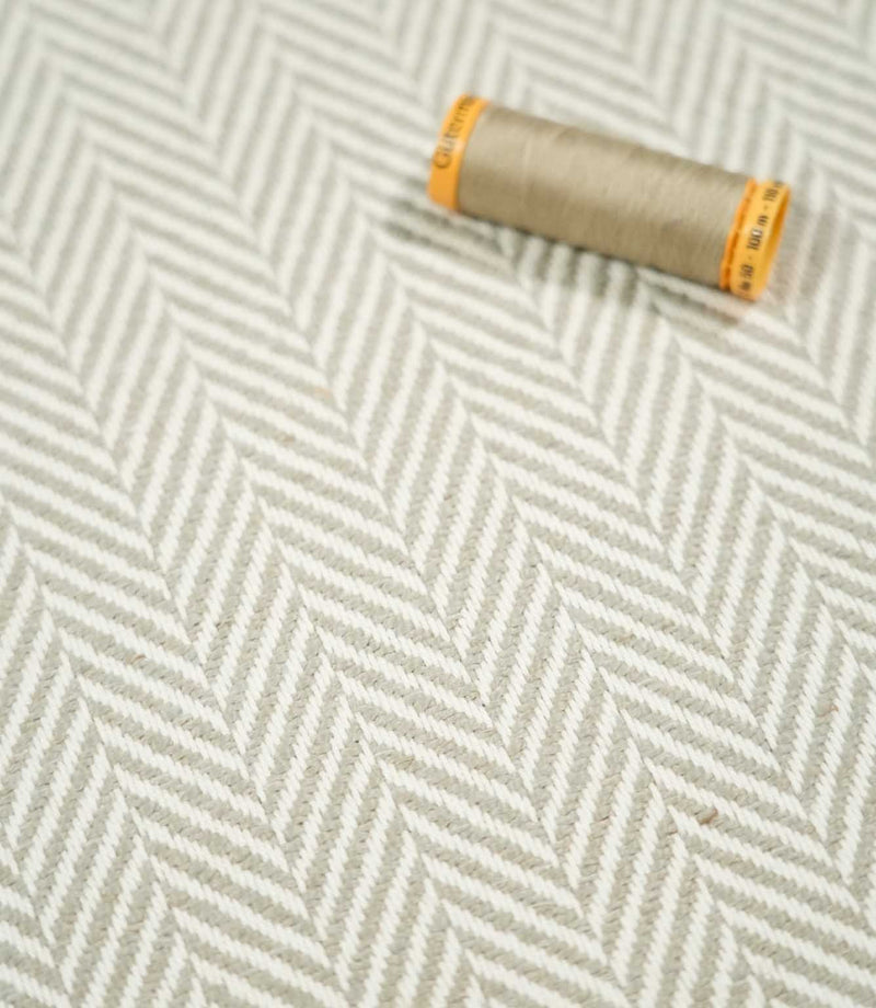 Neutral thread spool resting on Bampton Argent herringbone fabric, highlighting the coordinating pale grey-beige tones and off-white woven chevron pattern.
