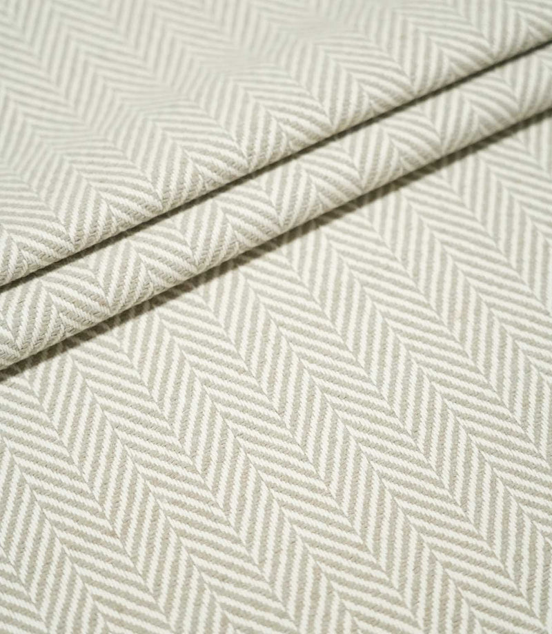 Layered folds of Bampton Argent herringbone fabric, highlighting the structured weave and repeating neutral grey-beige and off-white herringbone pattern.