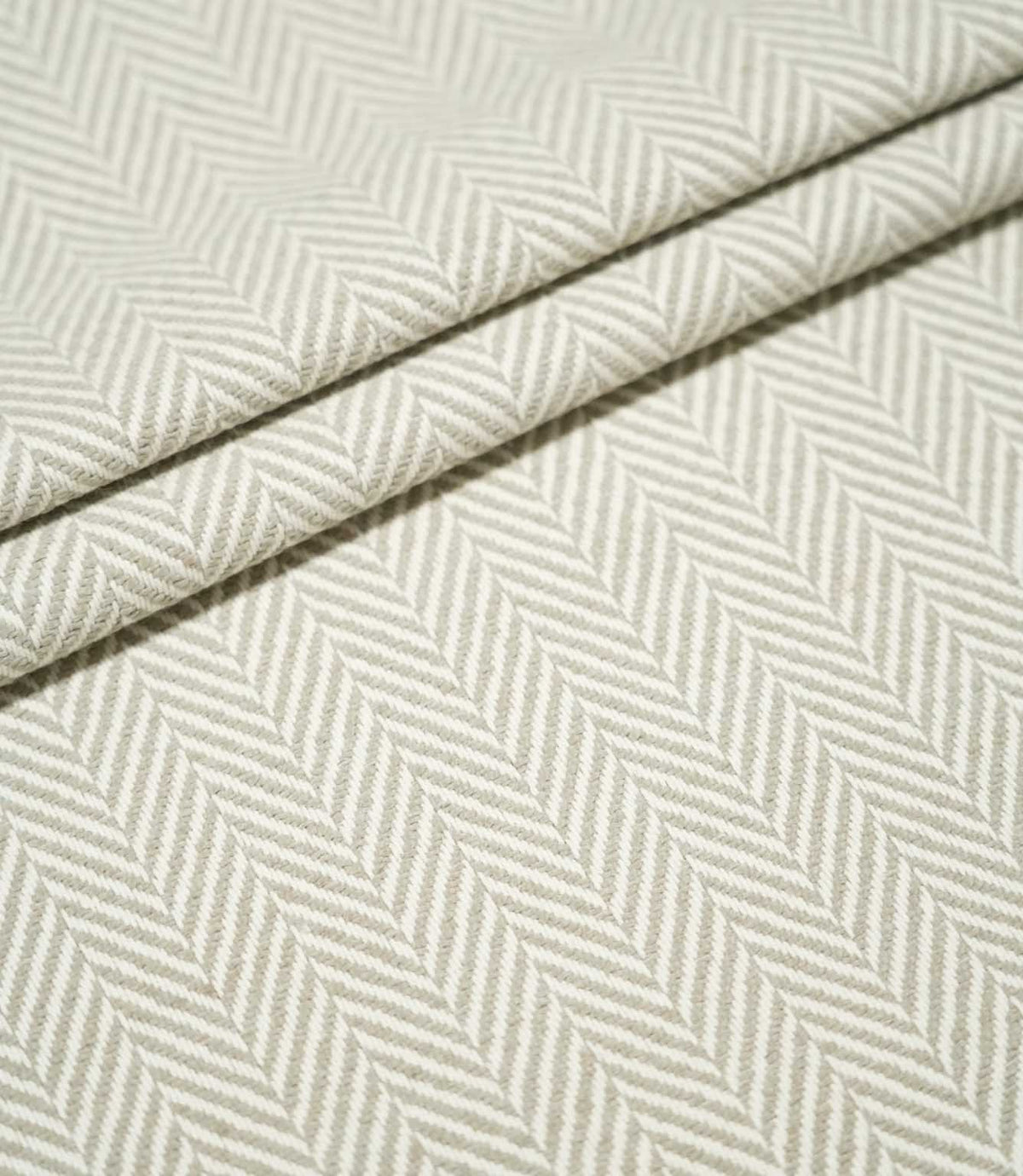 Layered folds of Bampton Argent herringbone fabric, highlighting the structured weave and repeating neutral grey-beige and off-white herringbone pattern.