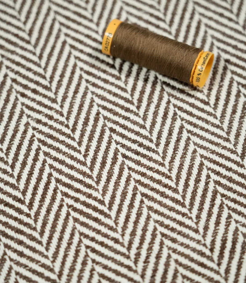Brown thread spool resting on Bampton Bison herringbone fabric, highlighting the coordinating colour and detailed brown and off-white chevron weave.