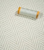 Pale grey thread spool resting on Bampton Elephant Grey herringbone fabric, highlighting the coordinating neutral tones and textured chevron weave.