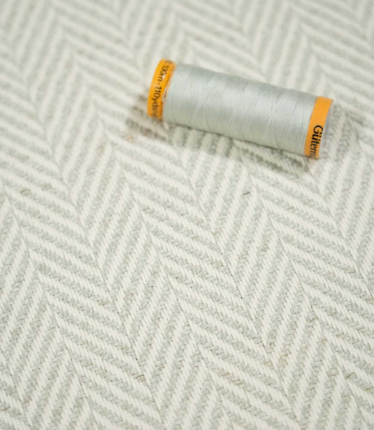 Pale grey thread spool resting on Bampton Elephant Grey herringbone fabric, highlighting the coordinating neutral tones and textured chevron weave.