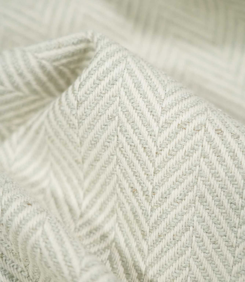 Macro close-up of Bampton Elephant Grey herringbone fabric draped into a soft fold, revealing the detailed pale grey and off-white weave, individual fibres and tactile texture.