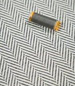 Grey thread spool resting on Bampton Grey herringbone fabric, showing the scale and fine woven detail of the grey and off-white chevron design.