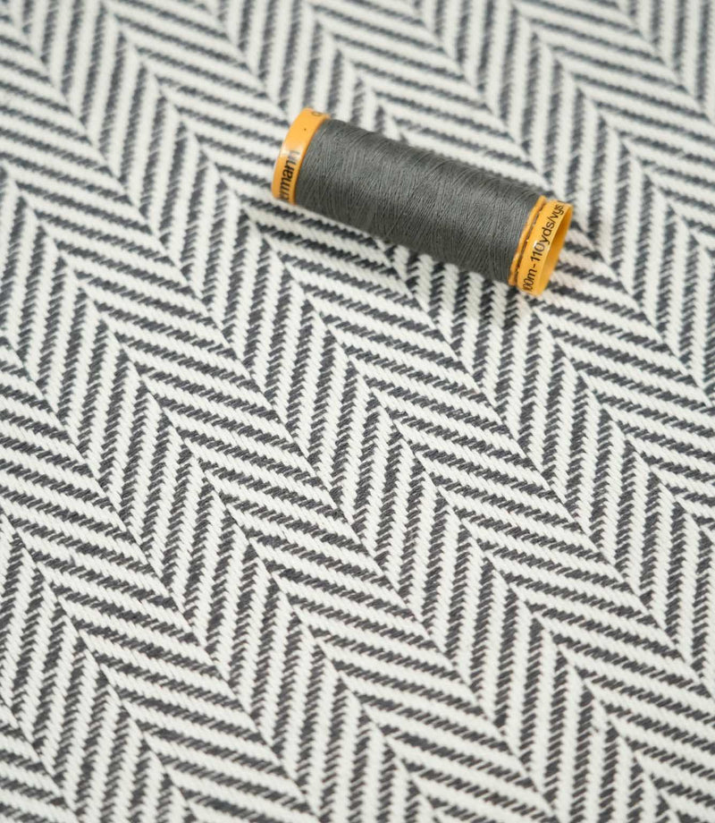 Grey thread spool resting on Bampton Grey herringbone fabric, showing the scale and fine woven detail of the grey and off-white chevron design.