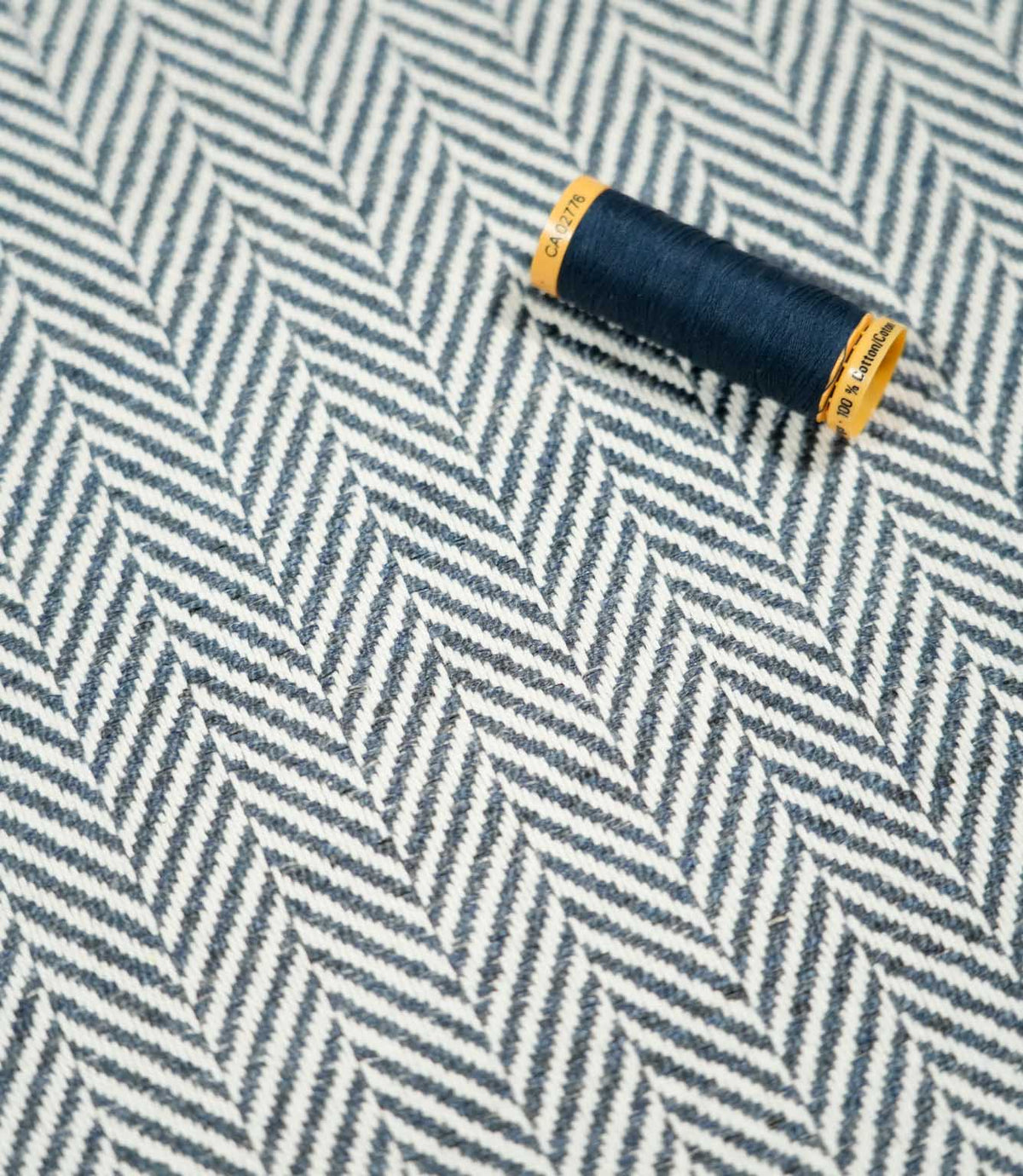 Navy thread spool resting on Bampton Ink herringbone fabric, highlighting the coordinating deep blue tones, off-white chevron pattern and tactile woven texture.