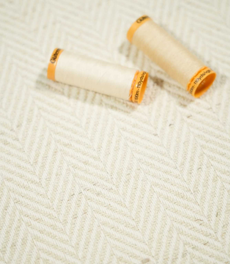 Two cream thread spools resting on Bampton Jasmine herringbone fabric, highlighting the warm neutral tone, subtle chevron design and woven texture.