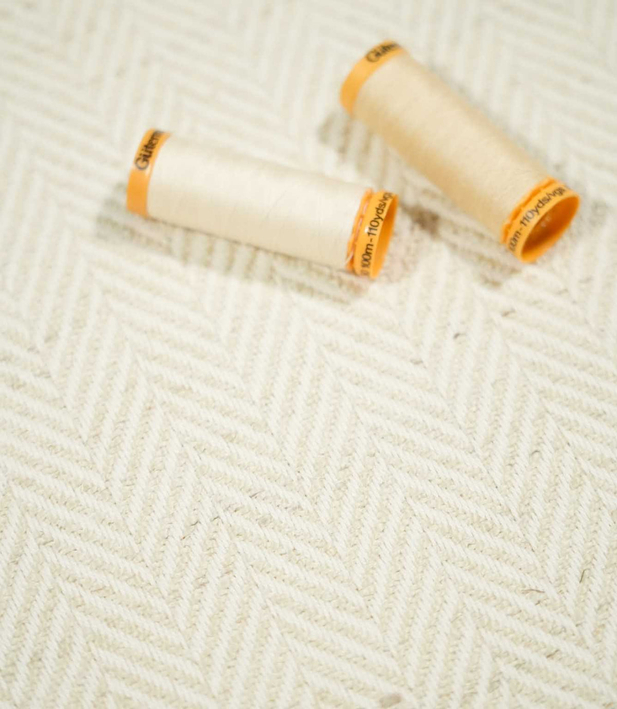 Two cream thread spools resting on Bampton Jasmine herringbone fabric, highlighting the warm neutral tone, subtle chevron design and woven texture.