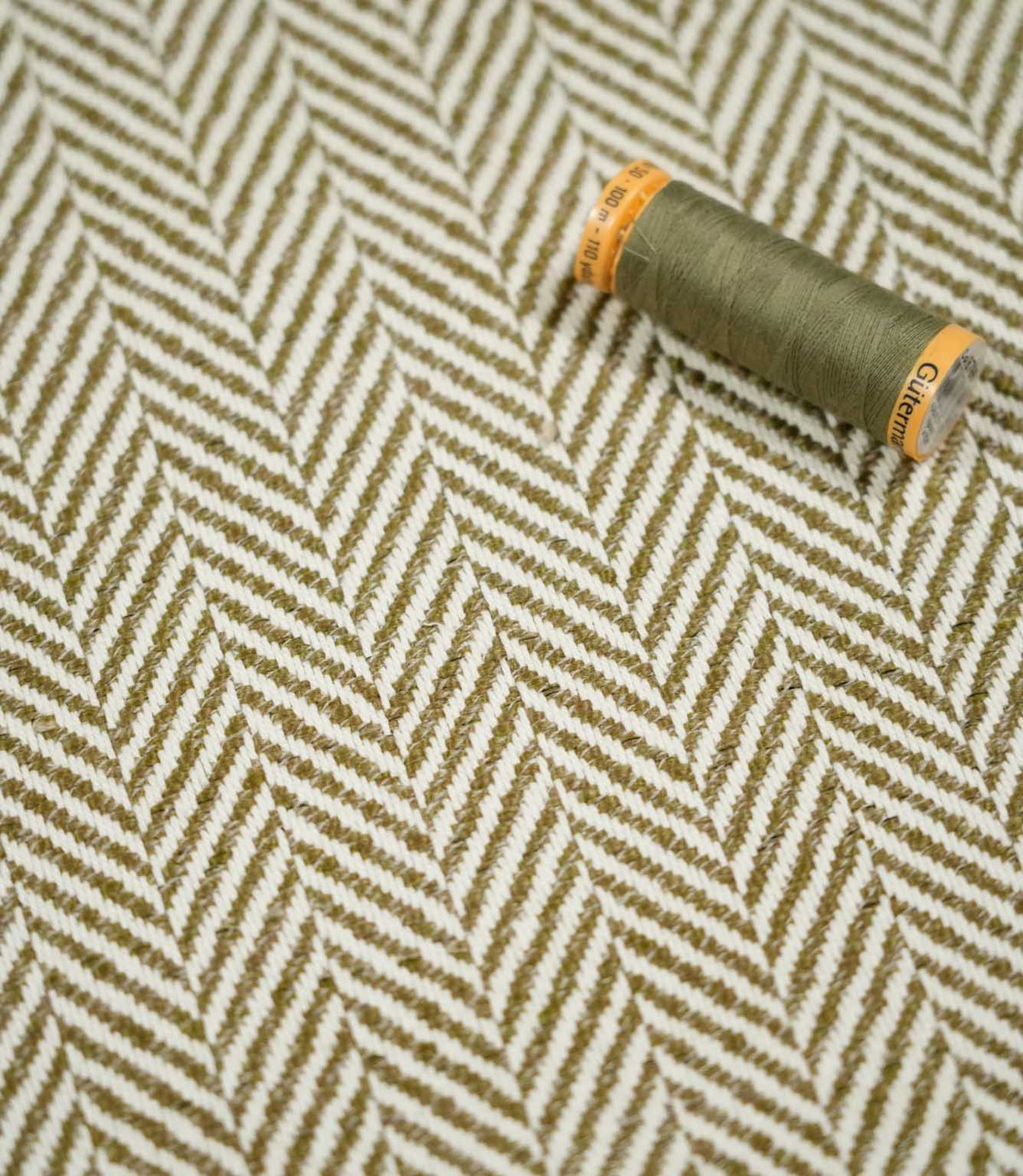 Olive green thread spool resting on Bampton Matcha herringbone fabric, highlighting the coordinating colour and detailed off-white and green chevron weave.