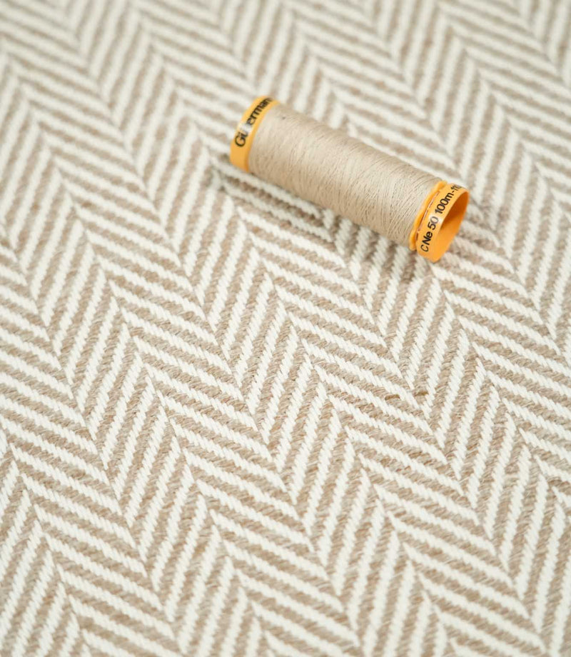 Taupe thread spool resting on Bampton Taupe herringbone fabric, highlighting the coordinating neutral tones and distinctive woven chevron pattern.