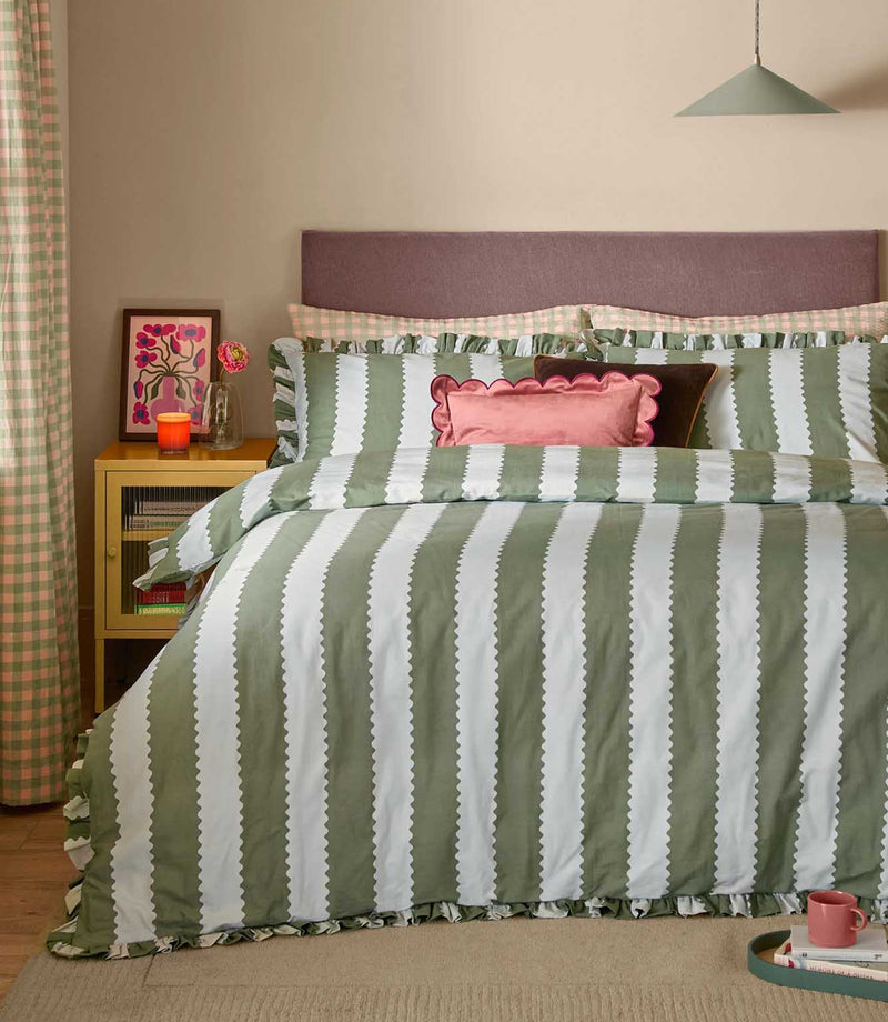 Birdie Stripe Ruffle duvet cover set in soft sage green and white, styled on a double bed with vertical scalloped stripes, ruffled edging, layered pink and brown cushions and coordinating checked curtains.