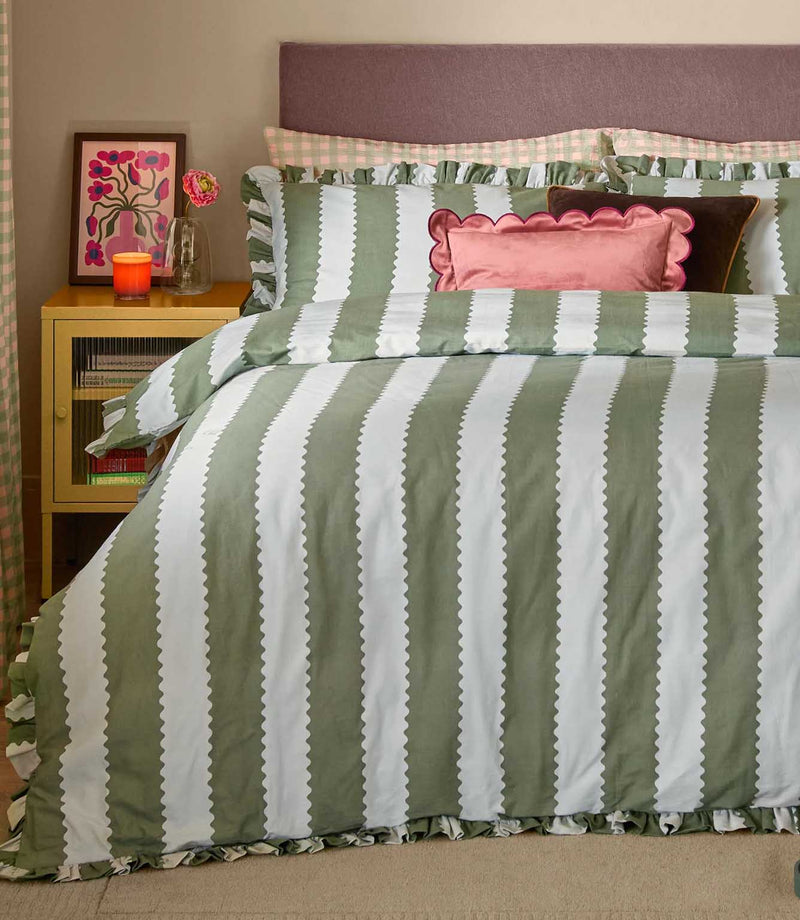 Front view of the Birdie Stripe Ruffle duvet cover set in sage green and white, featuring wide vertical scalloped stripes and gathered ruffle edging, styled with patterned pillows and pink accent cushions.