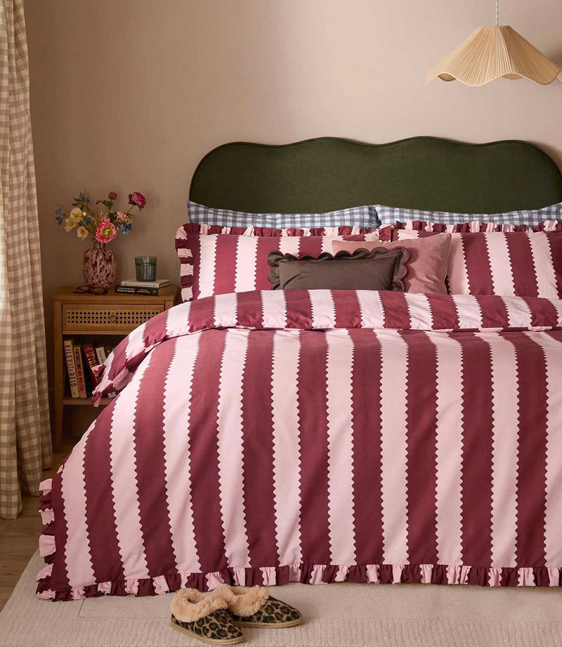 Birdie Stripe Ruffle duvet cover set in burgundy and soft blush pink, featuring wide scalloped vertical stripes and gathered ruffle edging, styled on a bed with coordinating cushions.